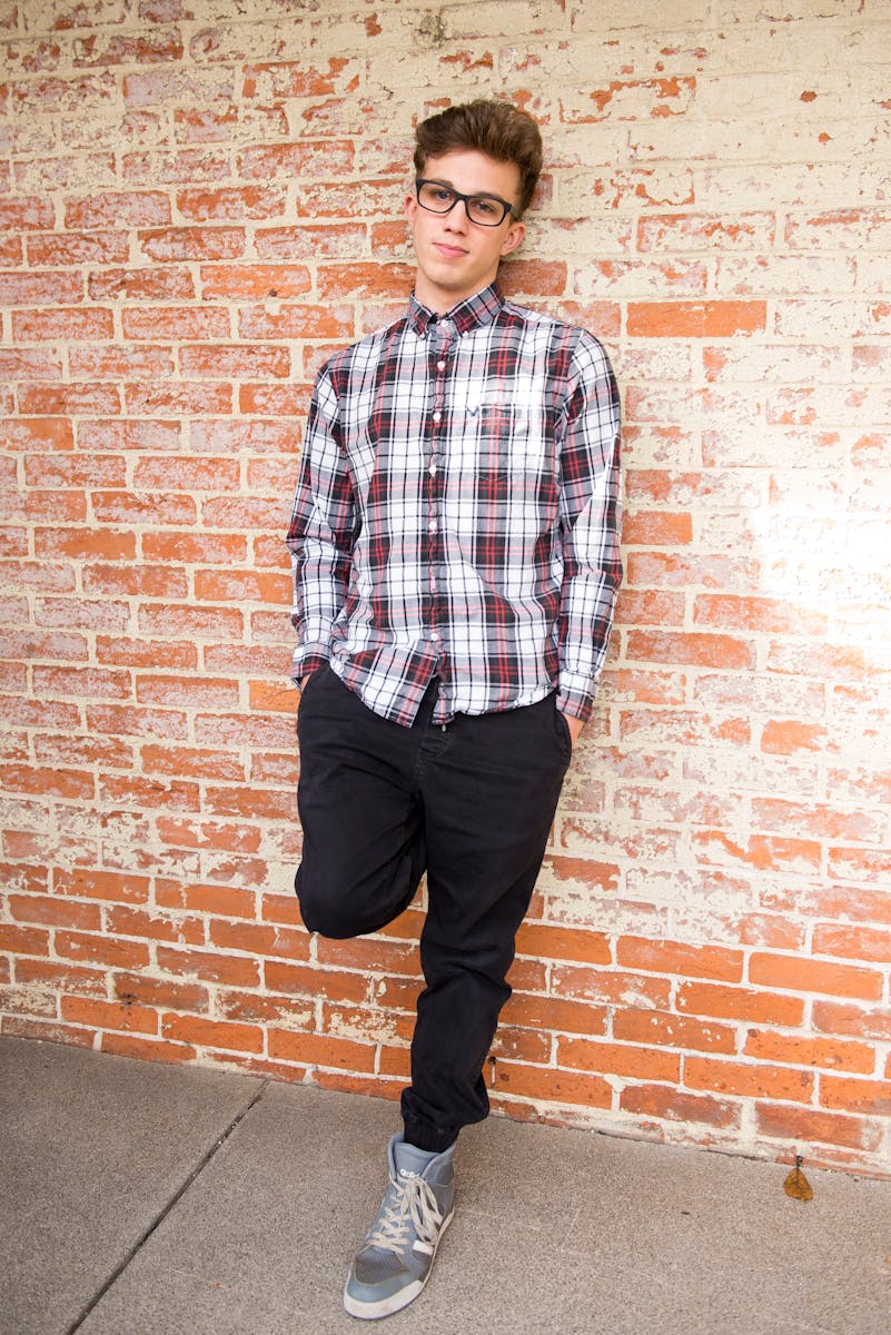 Teen boy in stylish attire posing against a brick wall outdoors.